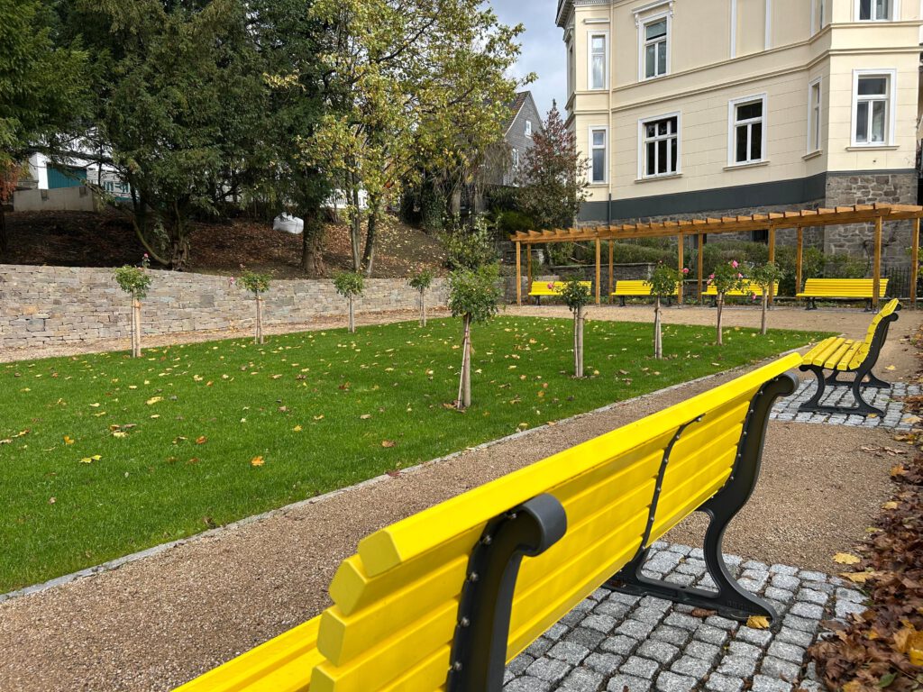Zwischenstand: gelbe Sitzbänke entlang eines säuberlich angelegten Wiesenstücks mit neu gepflanzten Rosenspaliersträuchern; am Ende der Wiese steht eine Pergola